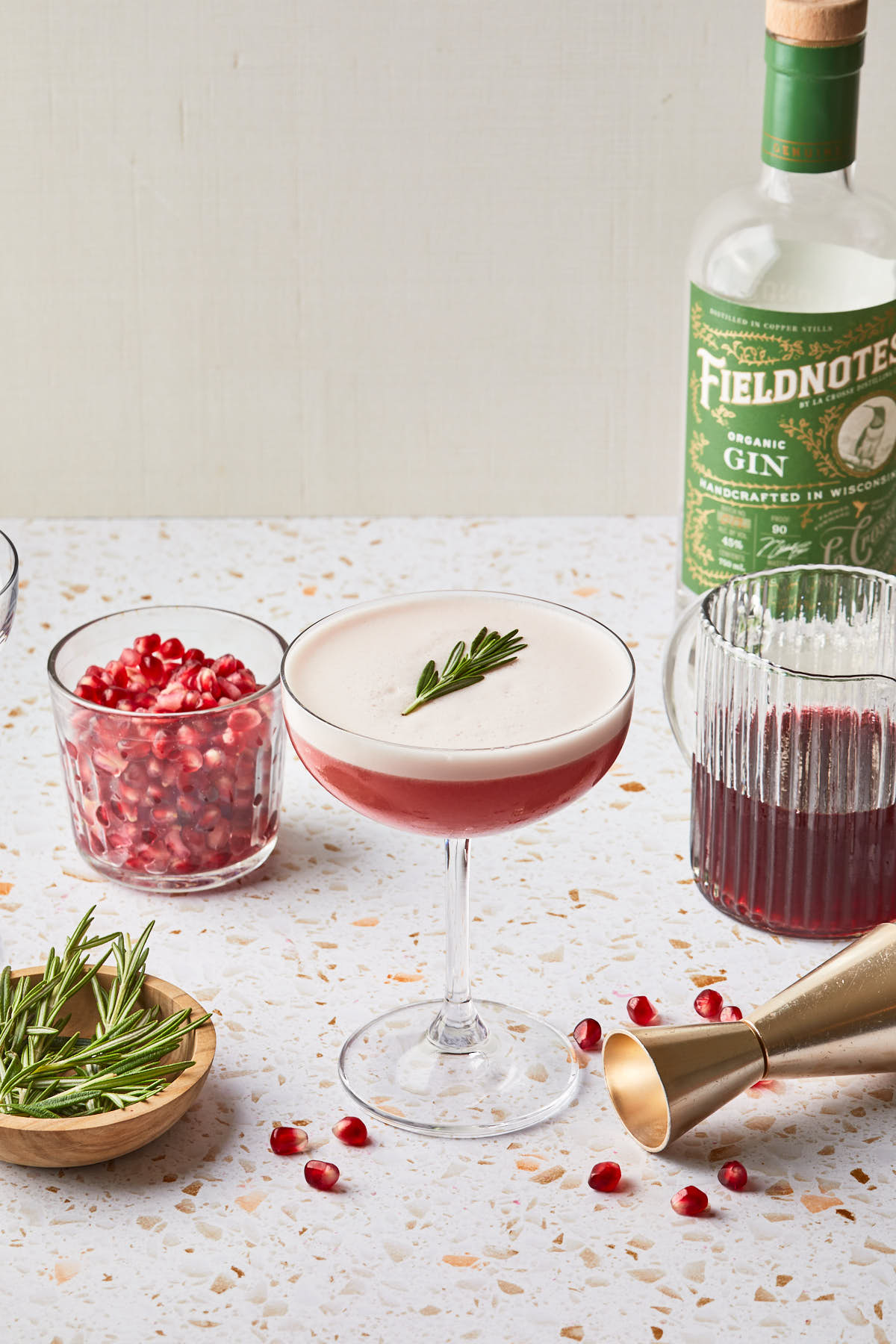 A coupe glass filled with a cocktail, topped with foam and a rosemary sprig next to a bowl of red seeds, a pitcher of red juice and a bottle of gin. 