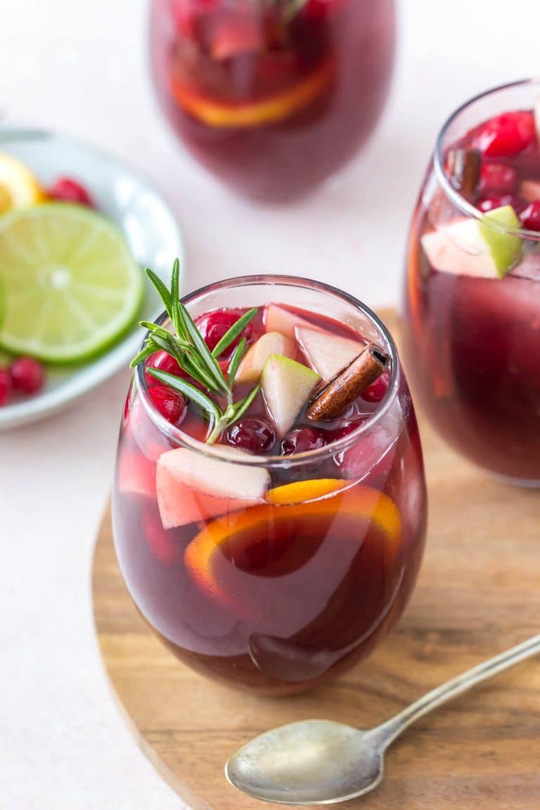 glass of red christmas sangria garnished with a rosemary sprig and cinnamon stick sitting on a wooden serving tray, two glasses and a small plate of fruit in the background