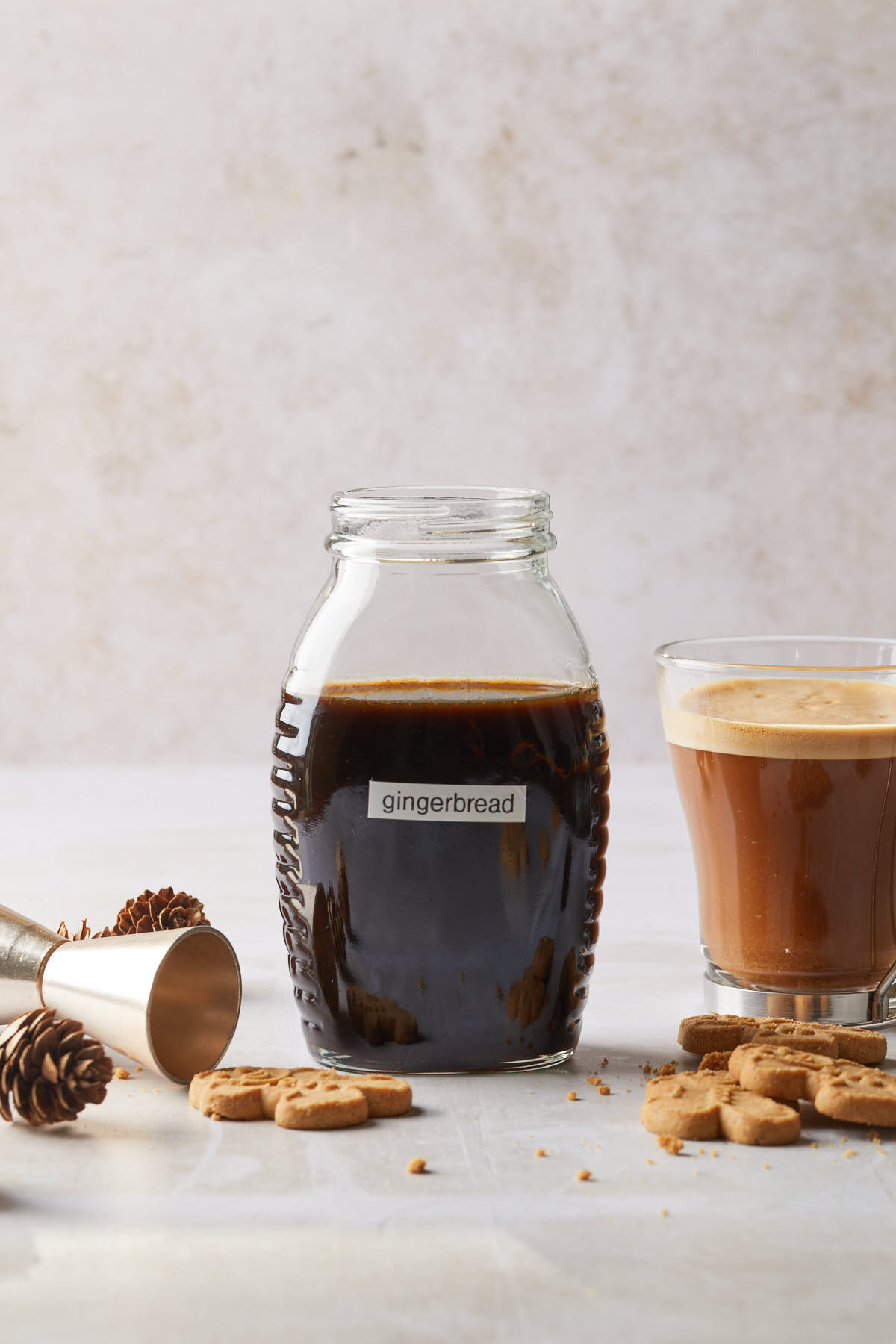 A bottle of gingerbread syrup with a glass of coffee, gingerbread cookies and a cocktail jigger. 