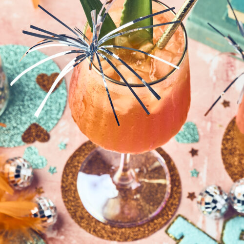 A festive orange showgirl spritz in a wine glass, garnished with pineapple leaves, a gold-striped straw, and shiny metallic streamers, sits on a decorated table with glitter, feathers, and small disco balls.