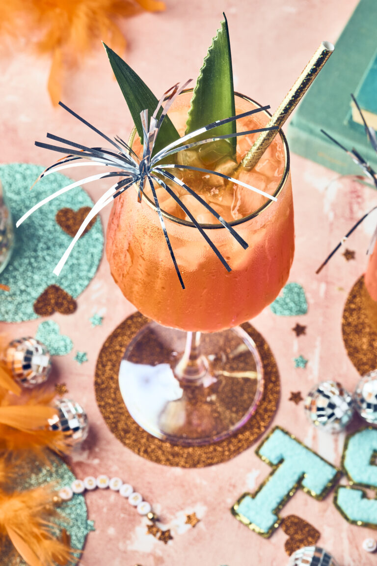 A festive orange showgirl spritz in a wine glass, garnished with pineapple leaves, a gold-striped straw, and shiny metallic streamers, sits on a decorated table with glitter, feathers, and small disco balls.