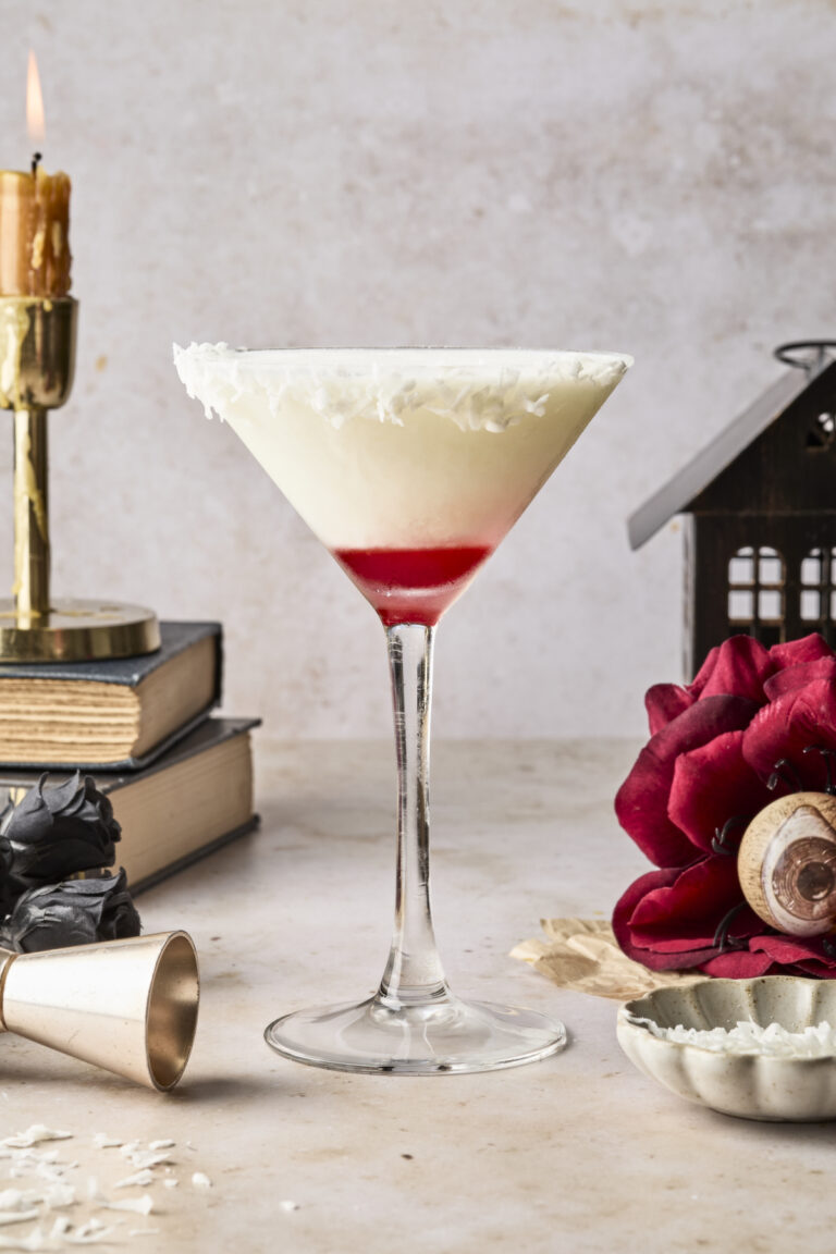 A white cocktail with a red layer at the bottom is served in a martini glass rimmed with coconut flakes. It sits on a light surface, with a red flower, books, a candle, and a small house decoration nearby.