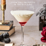A white and red layered cocktail in a martini glass with a coconut rim sits on a marble surface, surrounded by a candle, black roses, a book, a rose, and a cocktail jigger. Text reads, The Cocotini from Only Murders in the Building.
