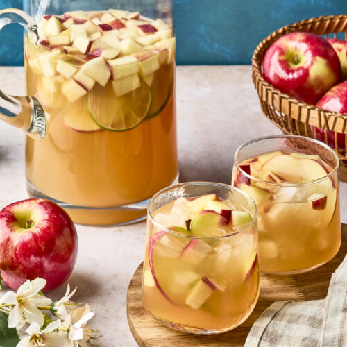 A glass pitcher and two glasses filled with apple and citrus fruit sangria sit on a table, surrounded by fresh red apples, a basket of apples, a beige napkin, and white blossoms, against a blue background.