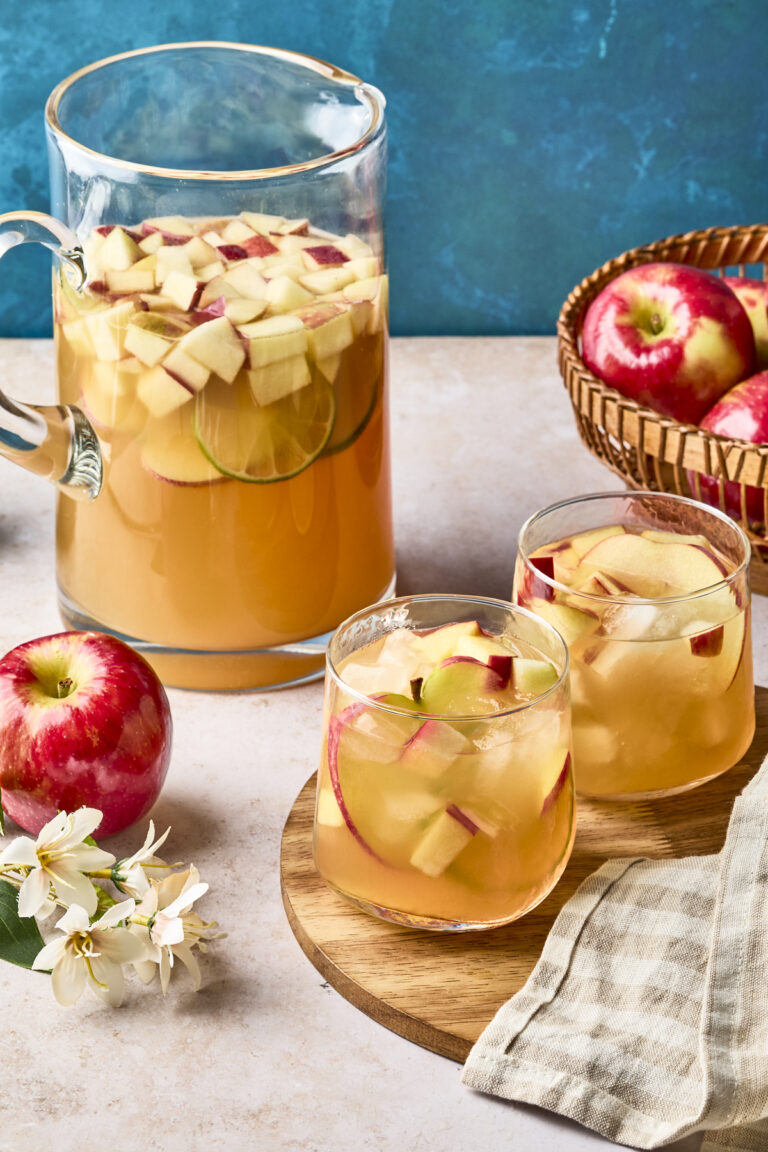 A glass pitcher and two glasses filled with apple and citrus fruit sangria sit on a table, surrounded by fresh red apples, a basket of apples, a beige napkin, and white blossoms, against a blue background.