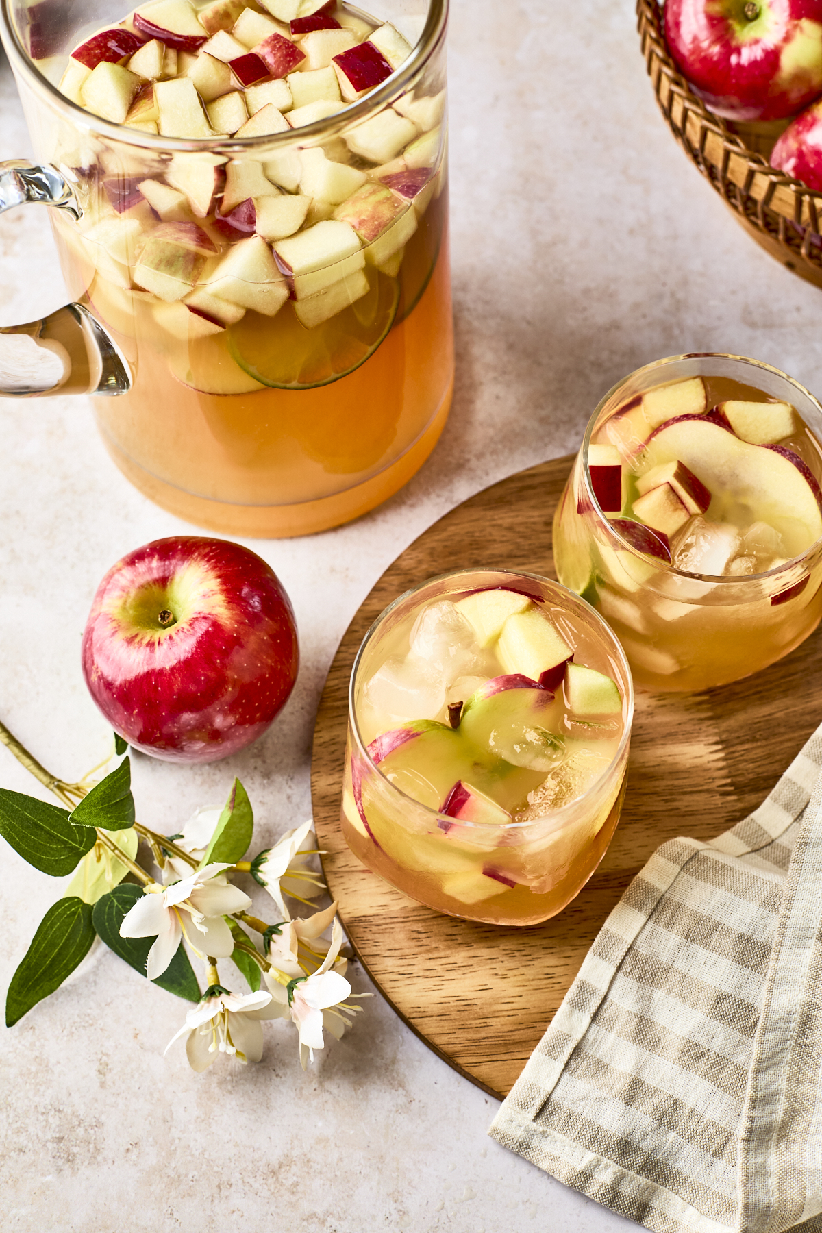 A pitcher and two glasses of apple sangria with ice and chopped red apple pieces, next to a whole red apple, white flowers, a striped napkin, and a basket of apples on a light surface.
